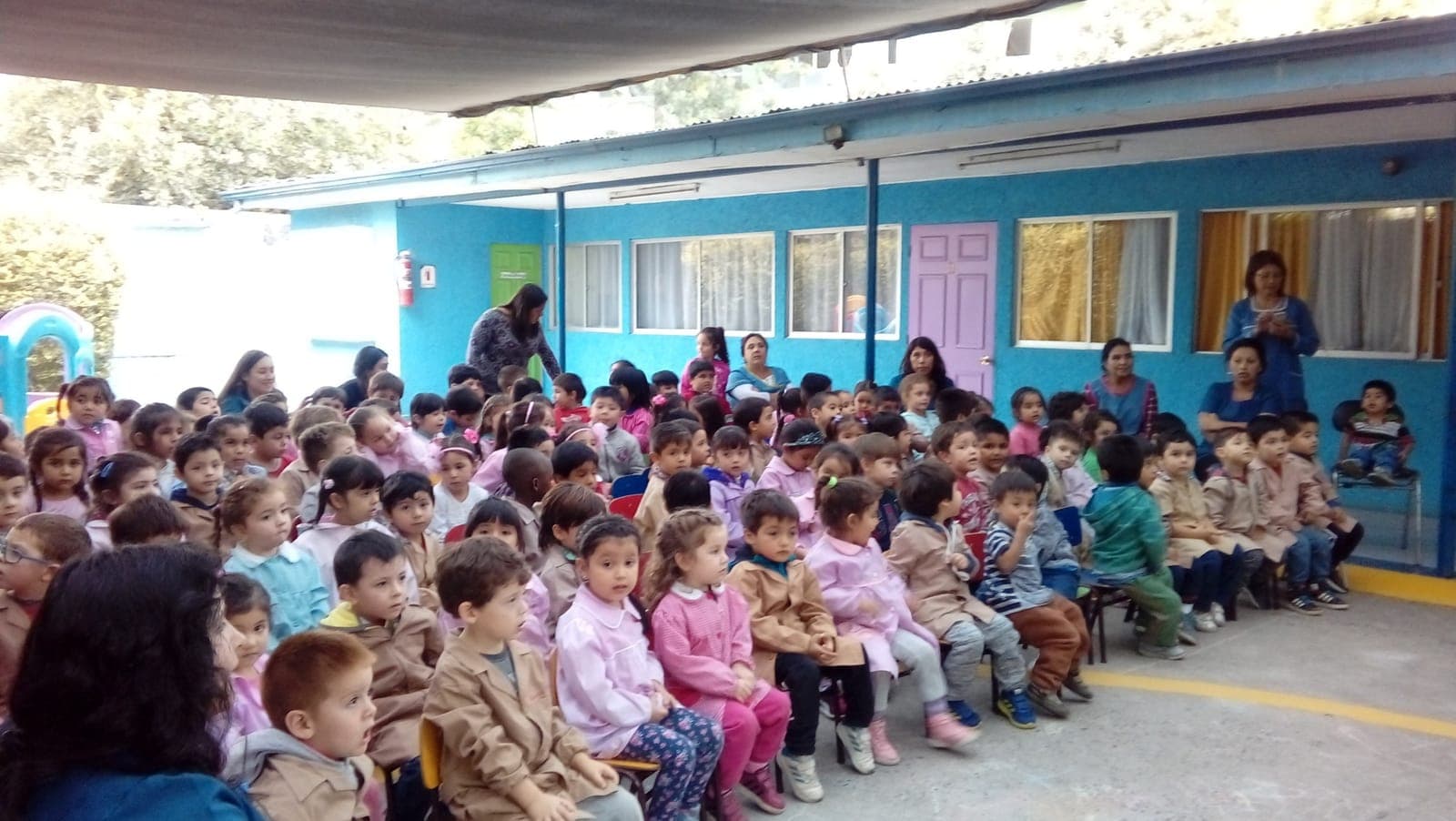 Niños y niñas de Kineret sentados en el patio del colegio, atentos a una presentación junto a sus tías.