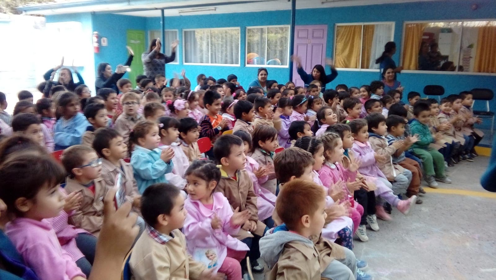 Niños y niñas con sus delantales aplauden sentados en el patio durante un acto del colegio.