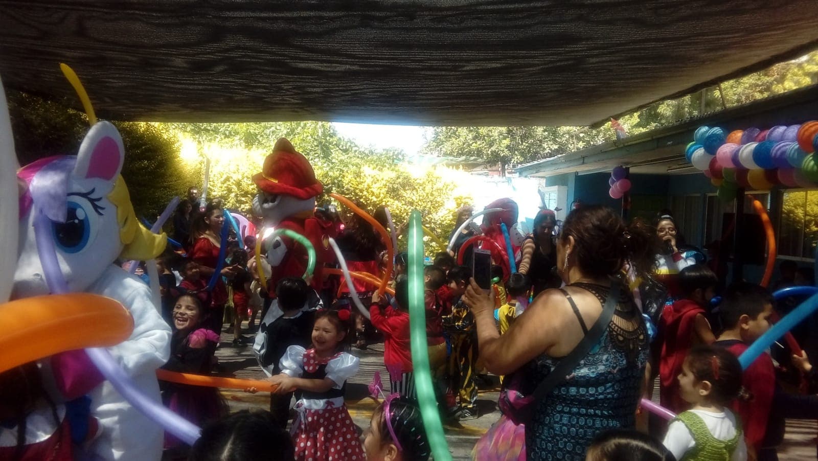 Niños disfrazados bailan con globos y mascotas en el patio mientras las familias toman fotos.