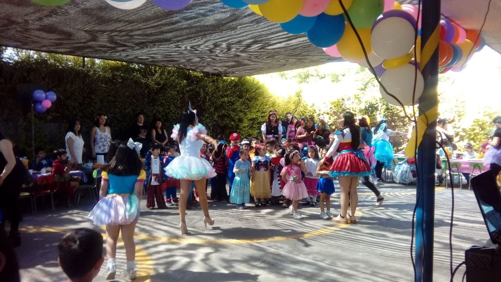 Animadoras disfrazadas bailan con niños y niñas disfrazados de princesas y superhéroes bajo guirnaldas de globos.