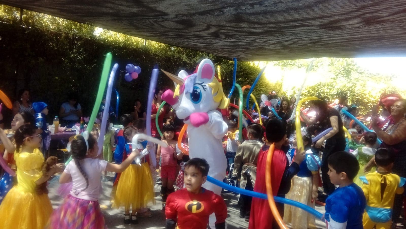 Niños y niñas disfrazados juegan con globos largos de colores junto a una mascota de unicornio.