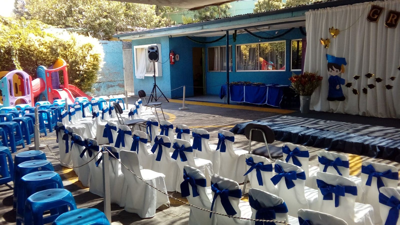 Patio preparado para la graduación, con sillas blancas decoradas con cintas azules frente al escenario.